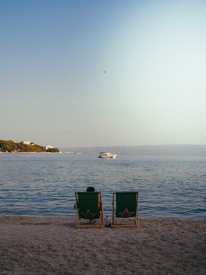 A person in a green deck chair on a pebble beach facing the sea